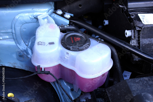 Close-up of translucent coolant expansion tank with black cap in a car engine bay, next to a purple liquid filler cap. Diesel engine cooling system.