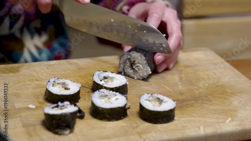 Female hands are cutting a sushi roll with nori algae seaweed and rice on a chopping board with a sharp knife. Different healthy ingredients are enclosed in the Japanese food roll. Healthy homemade fo
