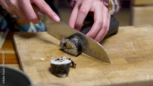 Female hands are cutting a sushi roll with nori algae seaweed and rice on a chopping board with a sharp knife. Different healthy ingredients are enclosed in the Japanese food roll. Healthy homemade fo