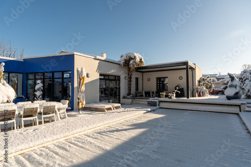 An elegant, modern house, in winter with a bioclimatic pergola providing shade for the outdoor area. The whole scene is covered in snow. 