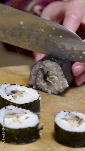 Homemade sushi roll gets cut with kitchen knife on wooden chopping board 2 vertical