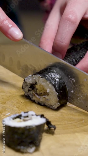 Homemade sushi roll gets cut with kitchen knife on wooden chopping board 1 vertical