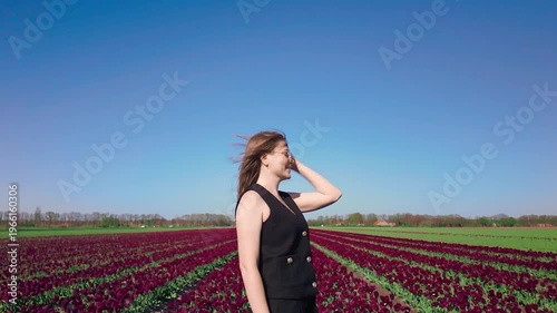 Woman stands confidently in vibrant tulip field under blue sky, calm posture and expression, outdoor fashion lifestyle, spring bloom environment.