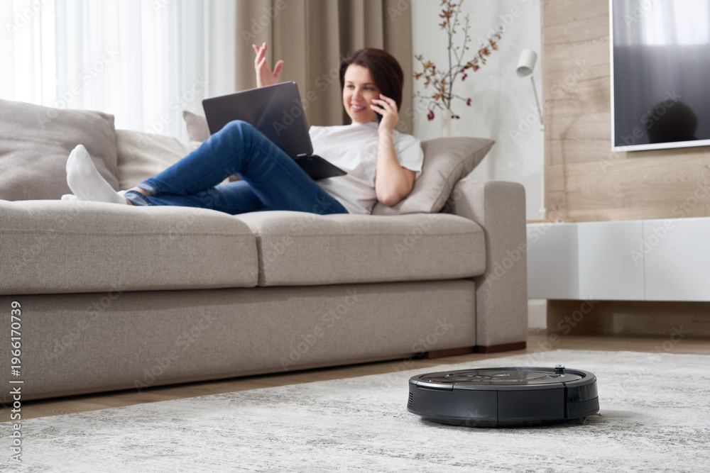 Fototapeta premium Robotic vacuum cleaner cleaning the room while woman resting on sofa