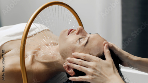 Female model receiving a relaxing hair treatment with water flowing from a circular device in a modern spa setting, soft lighting and minimalistic decor visible
