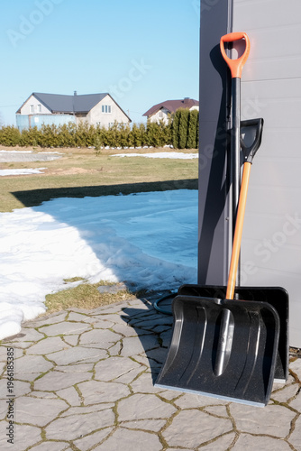 Two snow shovels leaning against a garage with melting snow, ready for winter cleanup. Residential scene highlighting snow removal tools and seasonal yard maintenance in early spring under blue sky.