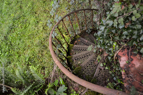 A rusty spiral metal staircase surrounded by dense greenery and overgrown plants in Busaco Park, Portugal. Themes explore decay, abandonment, and the return of nature to man-made structures.