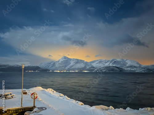 Mestervik, Norway - December 29, 2025: Malangen Brygger Resort. Glowing yellow sky-light on the horizon in the evening cloudscape shows mountain range covered in snow on opposite side of fjord. Hotel 