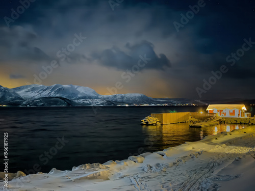 Mestervik, Norway - December 29, 2025: Malangen Brygger Resort. Glowing yellow sky-light on the horizon in the evening cloudscape shows mountain range covered in snow on opposite side of fjord. Hotel 