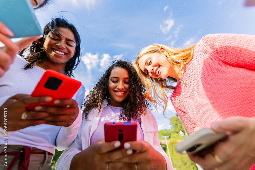 Obraz premium Close up of women using smartphones together outdoors. Diverse female friends connected through mobile technology and social media. Digital connection and online lifestyle concept.