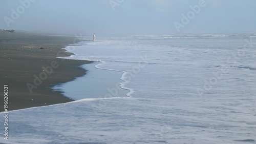 A lone person walks along the beach in Karekare, Auckland, New Zealand. The person is enjoying the solitude and beauty of the beach on a foggy day. Waves crash on the shore.