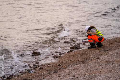 Bild auf Leinwand Female environmental scientist squats near shoreline, using smartphone and tablet to record water pollution data, highlighting environmental monitoring and coastal research
