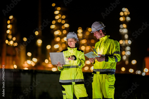Industrial teamwork concept. Male and female workers checking data on a computer during a night shift with beautiful glowing bokeh lights of a refinery behind them.