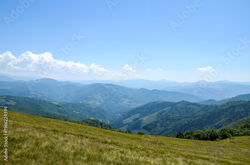 Panoramic view of green mountains and rolling hills covered in dense forest, with dramatic white cumulus clouds across a clear blue sky. Carpathian Mountains, Ukraine
