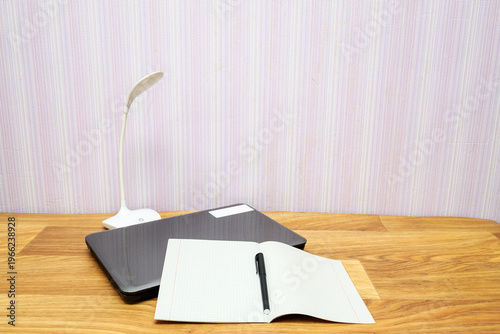 closed laptop, white lamp and open notebook on a wooden table in a bright interior