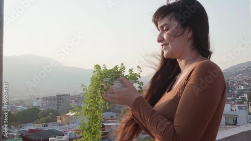 Young smiling woman holding a potted plant on a rooftop with her hair blowing in the wind, contemplating the urban landscape with mountains in the background at sunset