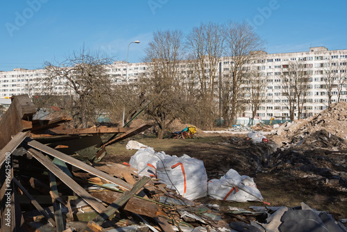 Construction waste and debris at a building demolition site in Narva, Estonia. Large bags and wood scraps against a background of residential multi-story houses in late afternoon.