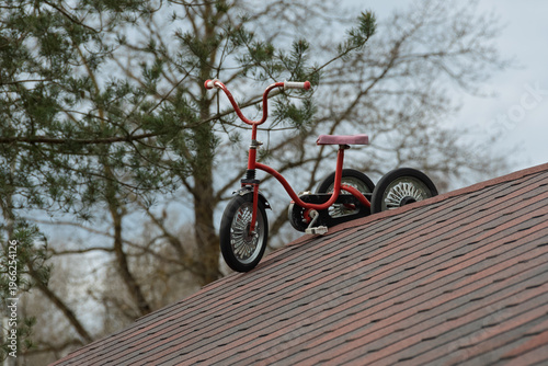 A red vintage children's tricycle standing on the steep shingle roof of a private house. A conceptual and surreal image against a background of spring trees in late April.