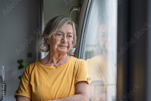Canvastavla An elderly woman in a yellow shirt stands by a window, arms crossed and gazing outside with a reflective, pensive expression