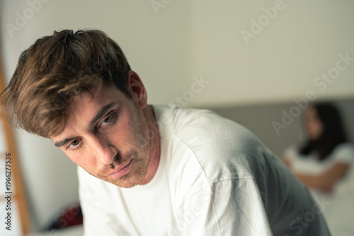 Bild auf Leinwand A young man sits on the edge of a bed looking withdrawn and pensive while his partner sits blurred in the background, conveying relationship tension, sadness, isolation and quiet reflection