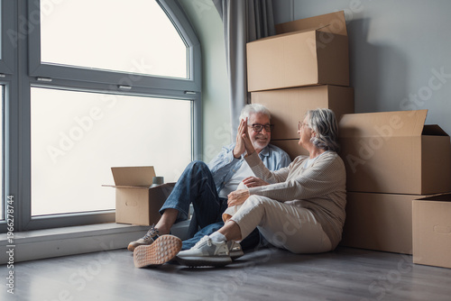 Fotografía Smiling senior couple sit on the floor among packed moving boxes by a large window, enjoying a quiet moment together as they settle into their new home during relocation