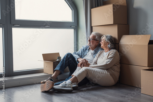 Photography A senior couple sits on the floor among packed boxes beside a large window, holding hands and smiling as they rest during moving day and look forward to settling into their new home