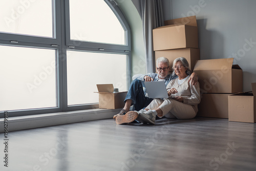 Fotografie A senior couple sits on the floor among packed boxes, using a laptop to organize moving details and share a quiet, affectionate moment together as they settle into their bright new apartment
