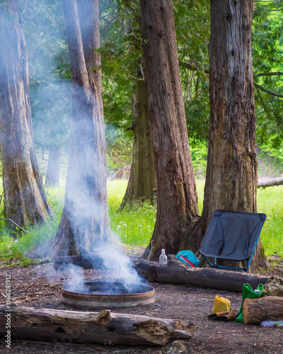 Smoke drifting from a fire ring in a backcountry camping site surrounded by fallen log benches and camping gear