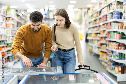 Bild auf Leinwand Couple shops in mega market and selects goods in fridge