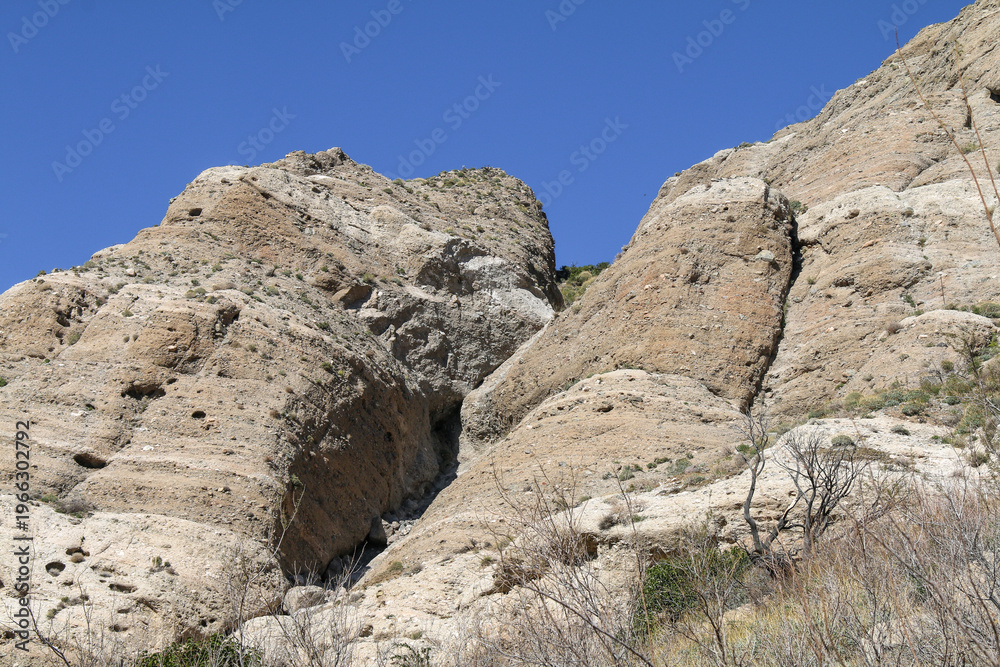 Fototapeta premium Large rugged tan rocks rise against a clear blue sky with sparse vegetation at their base creating a dramatic and dry mountainous landscape full of natural formations