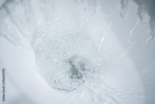 Water swirling in a white ceramic toilet bowl during a flush, foamy bubbles and motion blur forming a vortex around the drain, clean bathroom close up with bright highlights and minimal white interior