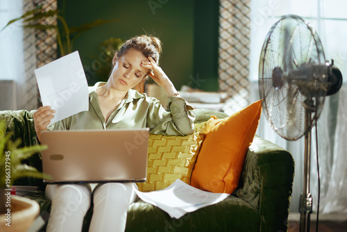 Fotografija A mature woman on a green velvet couch looks exhausted, fanning herself with paper while working on a laptop