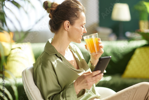 A woman enjoys a fresh glass of orange juice while browsing her smartphone during a peaceful morning routine in a sunlit living room.