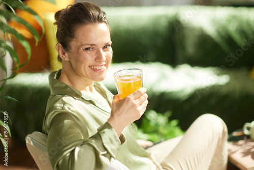 Tableau sur toile A smiling woman sits comfortably in her living room, enjoying a glass of orange juice as part of her healthy morning habits and relaxed home lifestyle