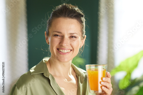 A cheerful middle-aged woman in a green shirt enjoys a glass of fresh orange juice during her healthy morning routine in a sunlit room filled with indoor plants.