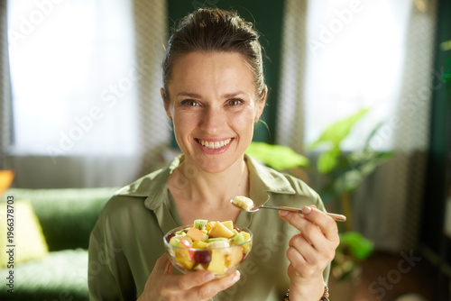 Happy woman holds a glass bowl of mixed fruit salad, savoring a spoonful of banana in her cozy, sunlit living room.
