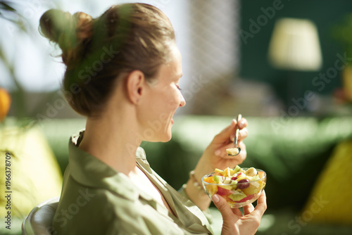 A woman in profile smiles while eating a mixed fruit salad in her living room. The cozy home interior and natural light highlight a moment of healthy snacking and self-care.