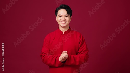 An Asian man stands relaxed against a red backdrop, cheerfully saying 