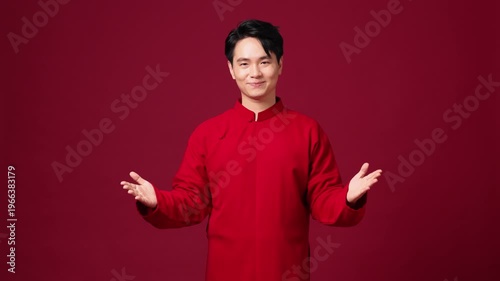 Happy Asian man presenting with both hands open, welcoming New Year mood, red backdrop.