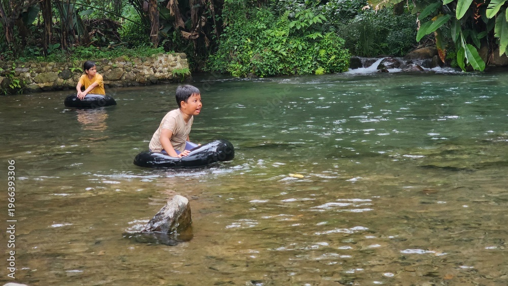 Fototapeta premium Two east asian kids ride a swim ring or swimming tyre and playing happily at a clean and shallow river. sstkTravel