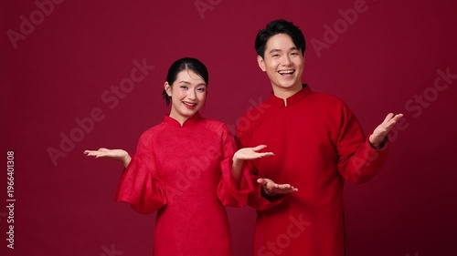 Asian couple presenting with open hands, red traditional outfits, friendly and welcoming New Year mood.