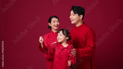 Asian parents clap along with their child, a heartwarming family moment against a vibrant red backdrop. The whole family looks in the same direction and smiles brightly