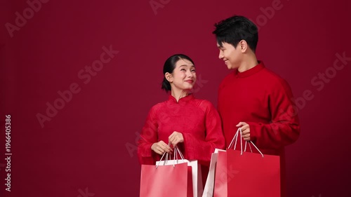 Asian couple holding shopping bags, cheerful pose on red background, New Year shopping concept.
