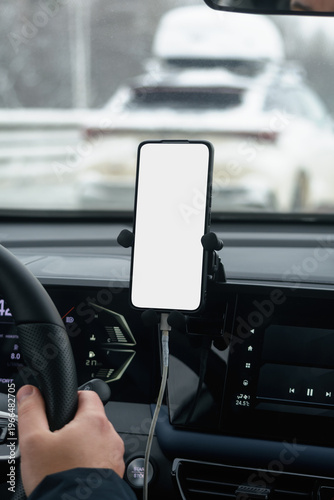 Blank smartphone mockup on dashboard mount inside modern car interior during winter drive with hand on steering wheel.