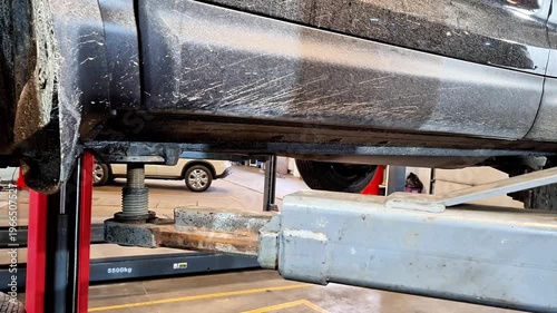 Underside view of vehicle elevated on hydraulic lift in automotive repair shop, showing lift arms supporting frame with background equipment, highlighting maintenance and inspection process