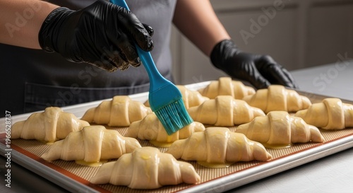 Professional baker wearing black gloves carefully brushes raw croissants with egg wash, preparing them for baking on a silicone mat