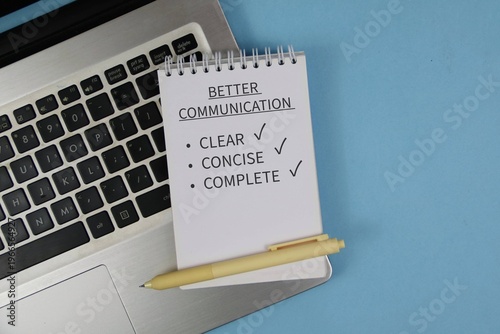 Notepad reading “Better Communication” with checklist items clear, concise and complete sits on a laptop keyboard beside a pen on blue background, conveying workplace communication and productivity.