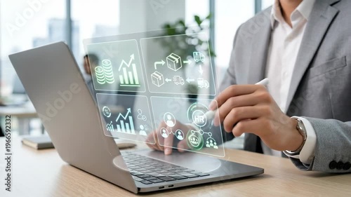 Professional businessman in a grey suit using a stylus and laptop to interact with a futuristic digital holographic interface showing supply chain logistics, financial charts, and data icons.