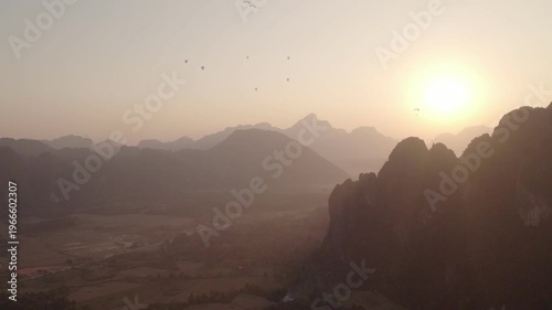 Very high altitude panning aerial view of mountainous landscape with paragliders flying around the sky and tropical rural landscape in the background.