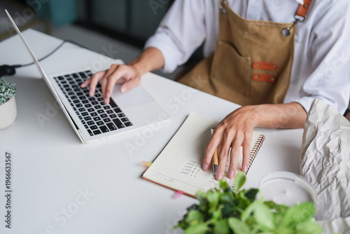 Asian Male Small Business Owner of Eco-Friendly Plant Shop Intently Managing Daily Sales, Income, and Expenses on Laptop.